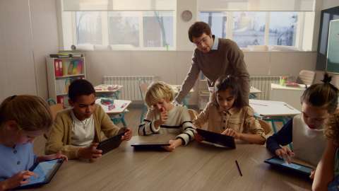 นักเรียนรุ่นเยาว์ 6 คนพร้อมแท็บเล็ตนั่งอยู่ที่โต๊ะที่ใช้ร่วมกันในห้องเรียน และผู้สอนมองงานที่ทำอยู่จากด้านหลังของนักเรียน