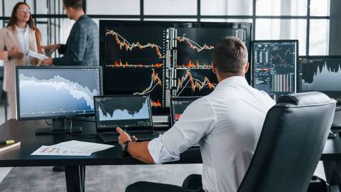 An analyst sits at a desk in front of multiple large-screen displays and laptops reviewing various trend lines and charts